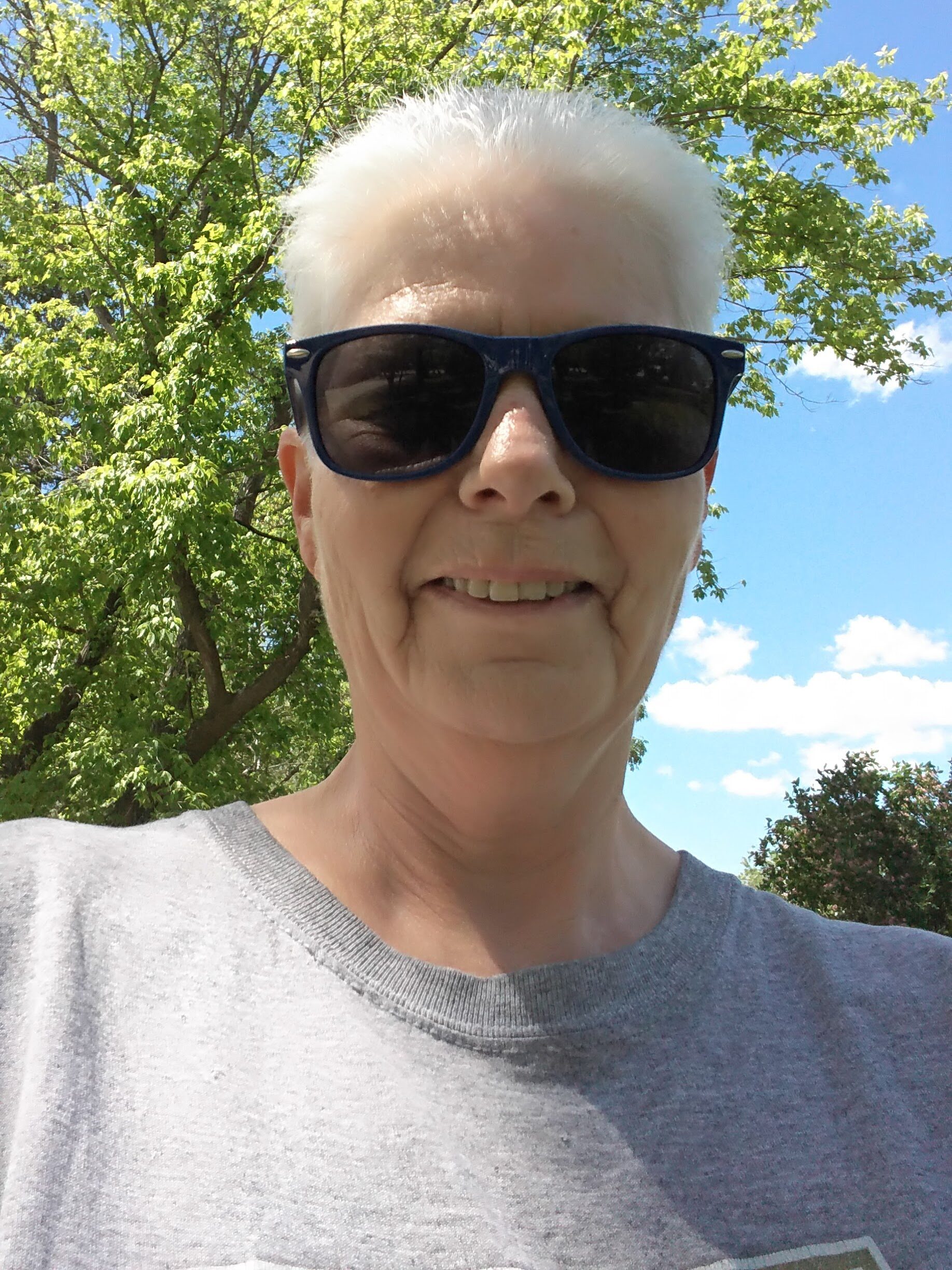 An outdoor pic of me wearing sunglasses standing in a park in front of trees and a blue sky