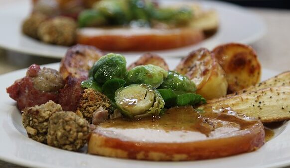 The image showcases a white plate filled with a holiday meal, including turkey and gravy, stuffing, and roasted potatoes, with Brussels sprouts prominently placed in the center. Behind this plate, another plate of Christmas dinner is softly visible in the faded background. The focus on the Brussels sprouts highlights their significance as a cherished family tradition in memory of the late husband, celebrating how they have become a beloved dish for the family, despite their mixed reputation among others.