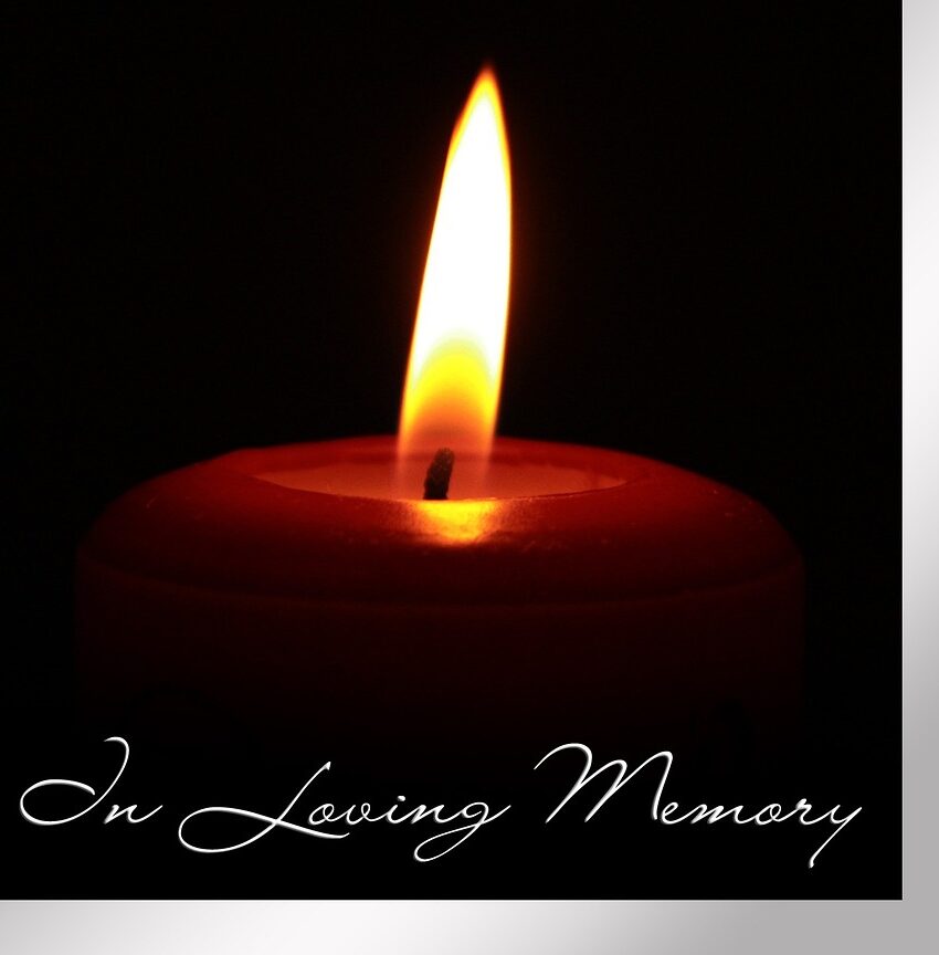 The image features a stubby lit candle, with only the rim visible as it emits a warm glow against a black background. Beneath the candle, the words 'In Loving Memory' are elegantly written in white. This serene image captures the essence of remembrance during the holiday season, reflecting the bittersweet emotions of honoring loved ones who are no longer present while navigating the festivities.